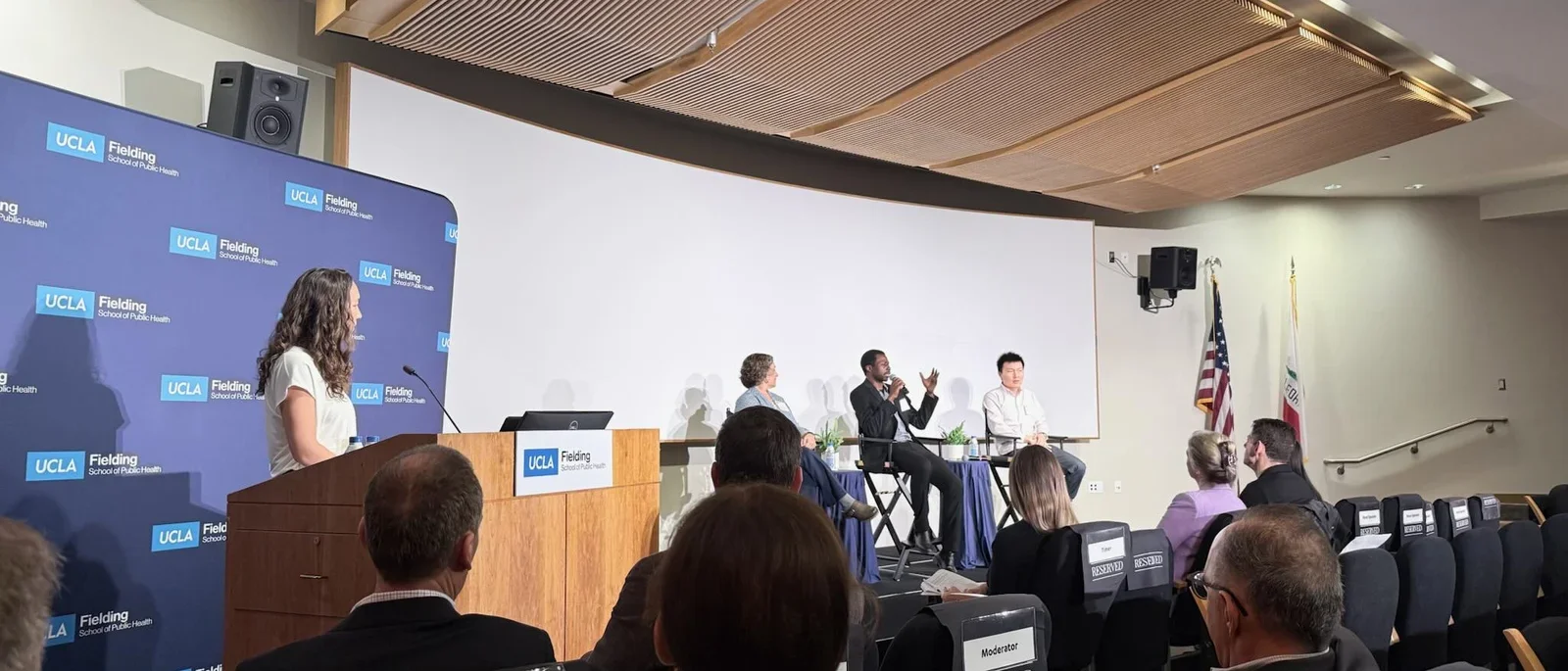 Woman at Podium with three panelists sitting on the stage