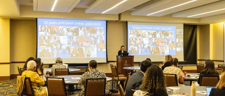Carol Mangione is at the podium with a picture quilt behind showing 20 years of RCMAR CHIME