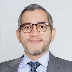 Man with glasses and grey hair and beard with a grey suite and blue tie and shirt
